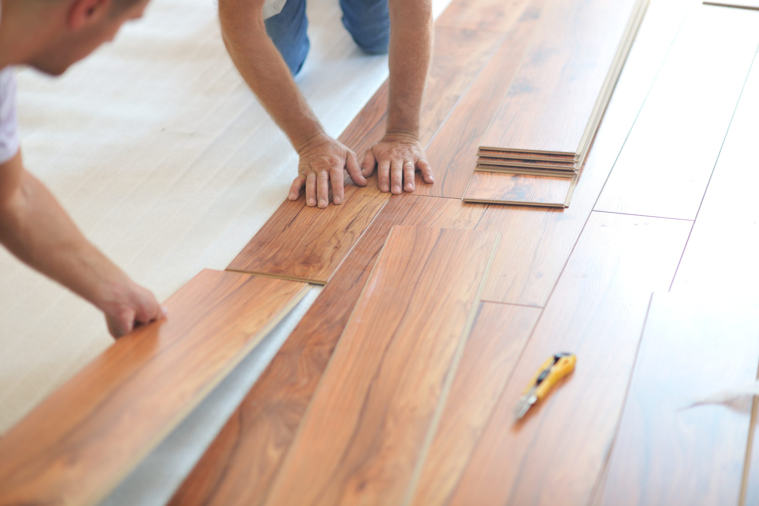  A professional flooring installer is installing laminate flooring in a new home. The installer is using a special saw to cut the laminate flooring to size. The installer is wearing safety glasses and a dust mask. The laminate flooring is a light brown color and has a wood grain pattern. The flooring is being installed in a living room.